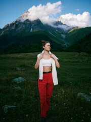 Fototapeta premium Woman in red pants and white top stands in alpine meadow with towering mountains, clouds and blue sky, enjoying outdoor scenery and nature tranquility.