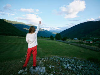 Fototapeta premium Woman standing on a rocky foreground, raising her hand toward a bright blue sky, with distant mountains and a green valley, conveying exploration, freedom, and outdoor adventure