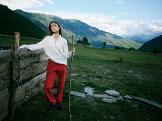 Woman leaning on a weathered wooden fence in a mountain valley, wearing a white knit sweater and red pants, holding a walking staff, gazing toward distant peaks under a clear blue sky. © SHOTPRIME STUDIO
