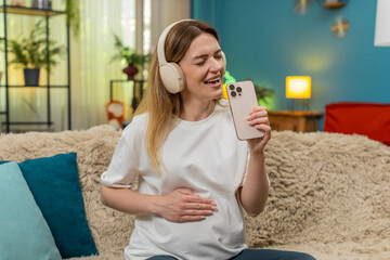 Pregnant middle-aged woman at home dances holding smartphone like microphone sings hit song full of joy. Caucasian girl on sofa moves feet shakes head laughs feels music so happy energy waiting baby.