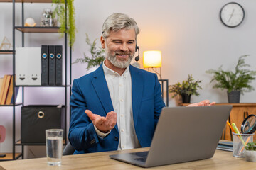 Middle-aged smiling businessman at home office wears headset assisting hotline caller via laptop. Freelancer guy at table gives rapid support ensuring customer satisfaction enhancing service quality