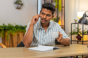 Indian man at home drinks coffee and writes goals in notebook ticks boxes and plans day with drive. Arabian guy at desk reviews list adds tasks sets priorities smiles feels focus and motivation aim