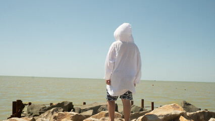 A teenager stands on a rocky seashore wearing a light hooded shirt and patterned swim shorts. He gazes out at the calm, sunlit sea under a clear summer sky, pensive and free.