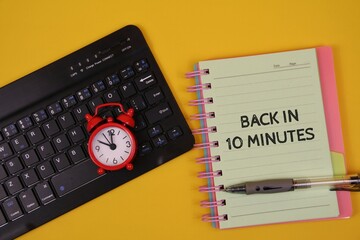 black keyboard, notebook reading 'Back In 10 Minutes', a red alarm clock, and a pen, suggesting focus, planning, and quick return to work. isolated on an yellow surface.