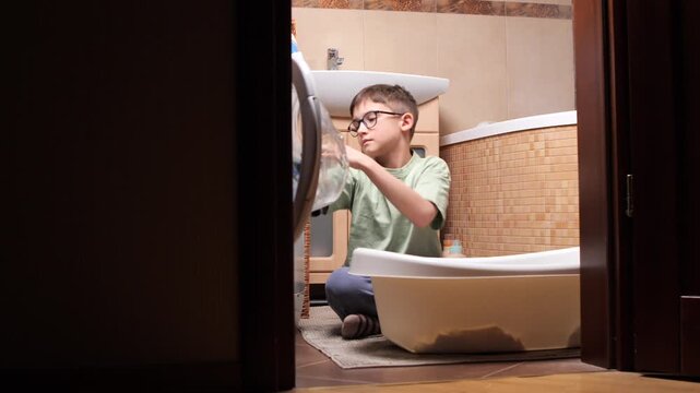Young boy learning household chores, carefully loading clothes from a basket into the front loading washing machine in a home bathroom, developing independence