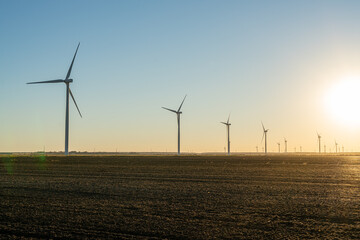 Wind turbines stand across open farmland at sunset, silhouetted against a warm golden sky as the sun drops below the horizon. © Eli