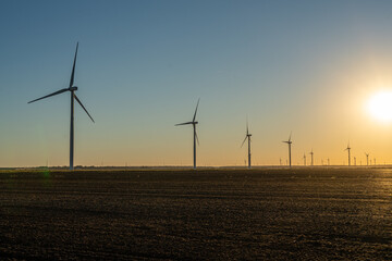 Wind turbines stand across open farmland at sunset, silhouetted against a warm golden sky as the sun drops below the horizon.