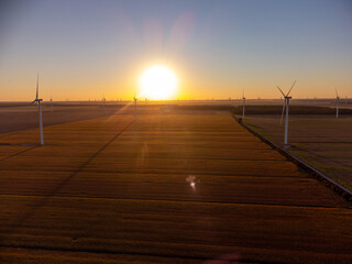 Aerial view of Wind turbines stand across open farmland as the sun sets on the horizon, casting...