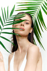 Fototapeta premium Elegant model with fresh makeup hiding behind palm leaves, embodying beauty and nature in a warm, tropical atmosphere, ideal for wellness themes.