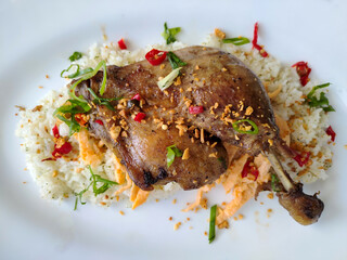 Fried duck leg served on seasoned rice with chili and herbs on a white plate, top view.