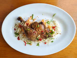 Fried duck leg served on seasoned rice with chili and herbs on a white plate, top view.