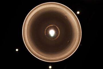 Close-up view from below of a minimalist ceiling light fixture with a bright, glowing bulb and a metallic reflector against a dark background, emphasizing modern interior design and illumination.