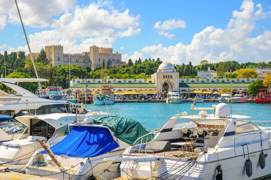 Boats docked at the Mandraki Harbour with the Nea Agora New Market and Palace of the Grand Masters of the Knights of Rhodes behind, in Rhodes, Greece.