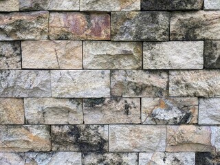 A close-up of a weathered, rustic stone wall texture, featuring rough-cut rectangular blocks in natural shades of tan, beige, and gray with dark mossy patches