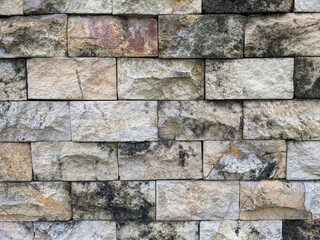 A close-up of a weathered, rustic stone wall texture, featuring rough-cut rectangular blocks in natural shades of tan, beige, and gray with dark mossy patches.