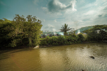River flowing through lush green tropical landscape with distant village and smoke rising into the sky