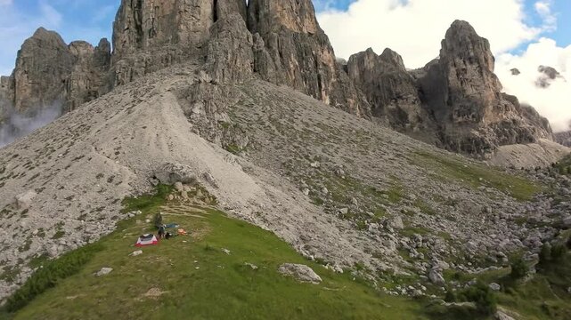 aerial alpine basecamp at rocky foothill; photographer documents small campsite against towering scree slope, clouds drifting, intimate outdoor scene with gear, guide planning next ascent