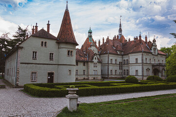 Historic European castle with towers, tiled roofs, and formal garden paths under a cloudy sky. Classic architecture and cultural heritage landmark photographed outdoors.

