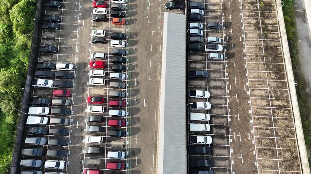 Aerial view taken with a drone of a large showroom parking area displaying neatly arranged new cars on both sides and a walkway in the middle.