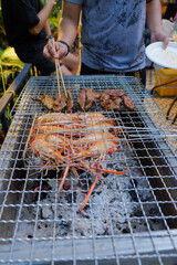 Person grilling large prawns and meat slices on a charcoal barbecue pit