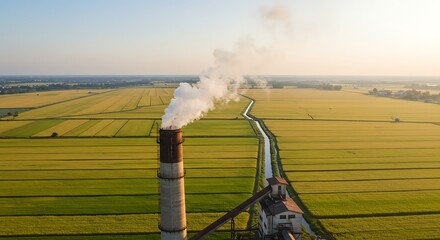Aerial view depicts factory smokestack emitting smoke over patchwork fields at sunset