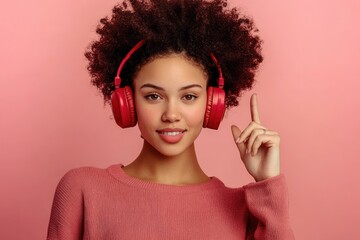 smiling young woman with curly hair wearing red headphones and matching sweater posing against pink background with finger raised in a positive gesture