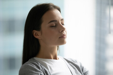 Inner peace. Close up shot tranquil beautiful young woman dressed in casual relax with closed eyes in light room taking deep breath of fresh air enjoy quiet meditation practice feel calm peace of mind