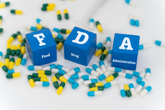 Three blue blocks labeled &lsquo;FDA&rsquo; (Food and Drug Administration) are positioned amid a dispersed collection of pharmaceutical capsules, symbolizing regulatory oversight