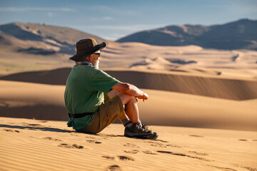 Senior man is sitting at sand dune and looking at sunset view. Desert vacations travel concept