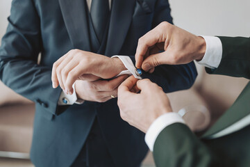 Grooming process before wedding ceremony as one man helps another with cufflink in a bright room in the morning