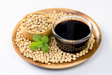 Soy sauce in a glass bowl and soybeans on a wooden plate against a white background.