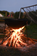 vertical photo. pot hangs above an open campfire, flames moving upward as the firewood crackles in the warm evening atmosphere. cooking cauldron sways slightly over a burning fire.