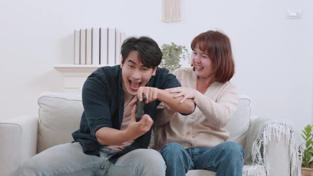 A lively Asian couple is seen close-up and full shot on a sofa, laughing and shouting with excitement while playing a video game using a wireless controller in their bright home.