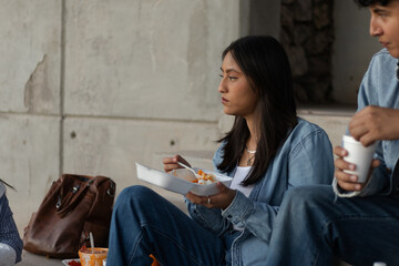 Young woman sitting outdoors on concrete steps, eating food from a takeout container, contemplating while sharing a casual street food meal with a friend during a break