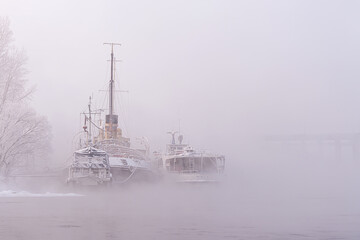 Atmospheric foggy landscape with nostalgic mood. Old decommissioned vessels, a Jung river tram and a tugboat or a river icebreaker permanently parked on the Yenisei River, shrouded in mystery and fog