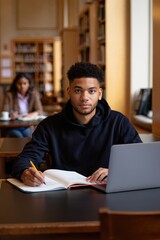 Fototapeta premium Approachable American college student studying in library with laptop and notebook