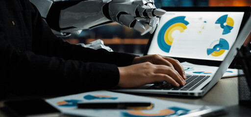 A robotic arm assists a human typing on a laptop in a modern office setting, showcasing a collaborative workspace enriched with vibrant graphic designs.Noogenesis