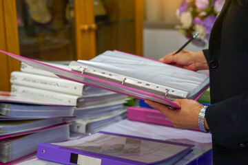 A person in a business suit is holding and reviewing a pink binder filled with documents, with a pencil in hand, surrounded by stacks of organized files on a table.