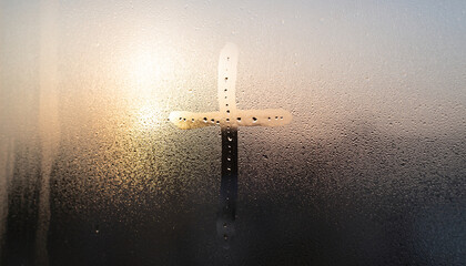 A Christian cross drawn on a steamy window with sunlight in the background.