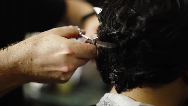 Close-up of a barber using scissors to trim the back of a client's dark wavy hair, with precise snips around the nape and neckline during a salon haircut and grooming session.