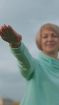 Senior woman extending palm for balance, teal sweatshirt and short hair smiling, blurred torso and cloudy sky backdrop, slow steady movement, wellness practice, gentle exercise session, encouraging