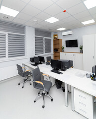 In a bright office space, several desks are arranged with computers. Windows provide natural light, and storage units are neatly placed. It is daytime and the area is tidy