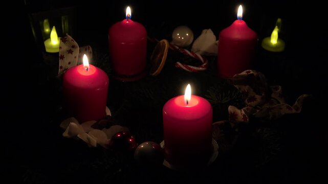 Ein Adventskranz mit vier brennenden roten Kerzen und zwei flackernden Laternen dahinter im Dunkeln