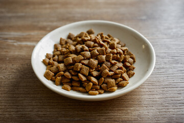 Dry pet food in a small white bowl placed on a wooden table in a kitchen setting during daylight hours