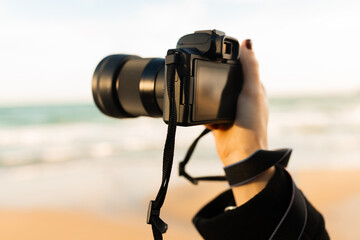 Close-up of woman holding a professional camera on the beach during golden hour, focusing on the lens and capturing outdoor photography moments.