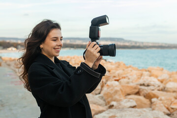 Young female photographer taking pictures outdoors by the sea, smiling while holding a professional camera with flash, capturing moments on a rocky coastline at sunset.