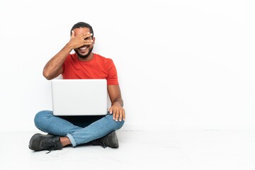 Young Ecuadorian man with a laptop sitting on the floor isolated on white background covering eyes by hands and smiling