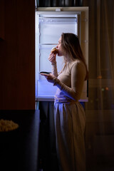 Female eating muffin while standing in front of open refrigerator