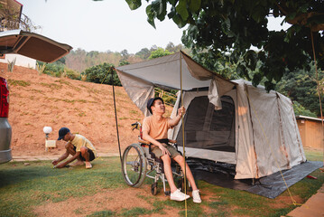A wheelchair user helps adjust a tent rope while camping outdoors as a companion sets up nearby. The scene shows accessible camping, teamwork, inclusion, and an active lifestyle surrounded by nature.