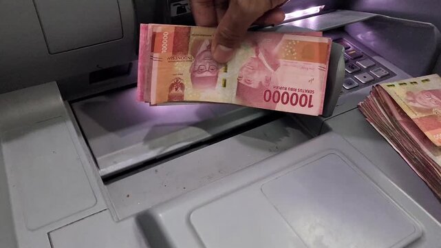 An Asian man is taking money out of a top-loading ATM machine that can be used for deposits and withdrawals in 100 thousand rupiah denominations, then counting it.